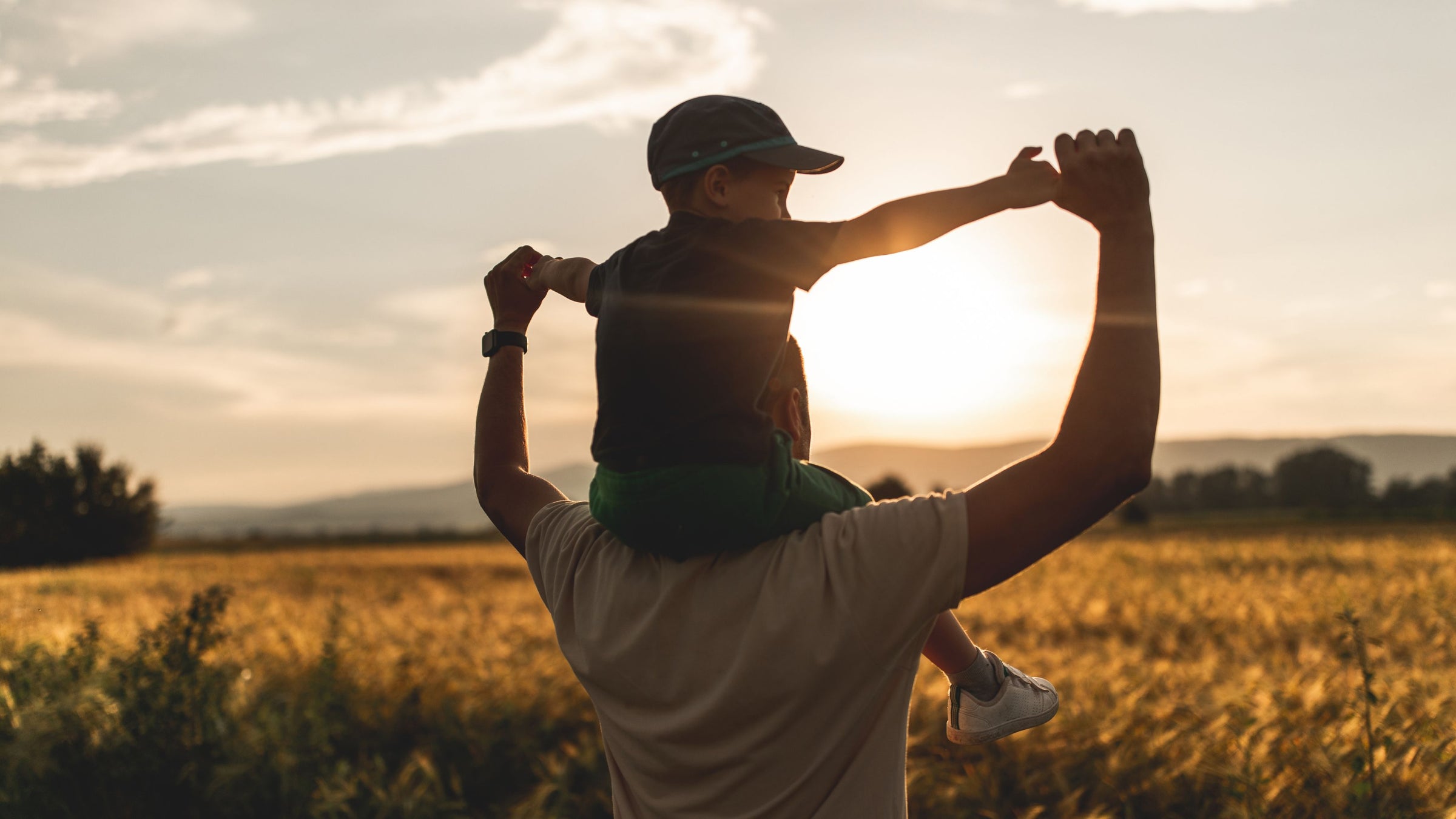 child on man's shoulders staring a field during sunset