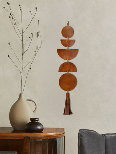 A modern, minimalist interior featuring a leather wall hanging composed of geometric shapes in warm brown tones with a tassel at the bottom. The piece hangs against a soft beige wall. Below, a wooden console holds a neutral-toned ceramic pitcher, a small black vase, and a dried botanical arrangement, adding an organic touch. A dark gray cushion is partially visible in the foreground.