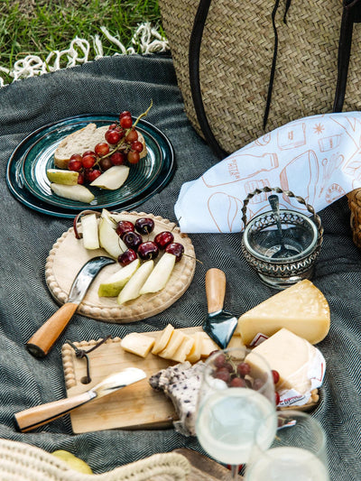 Picnic setup with a wooden cheese board, fresh fruit, and artisanal kitchen tools, featuring handcrafted knives and serving utensils from Joffa.