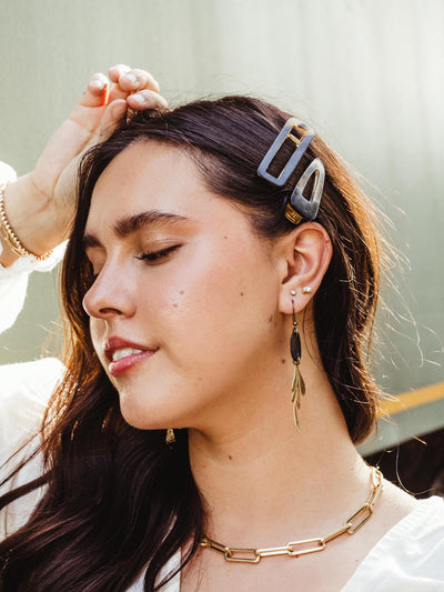 Woman wearing two geometric hair clips, gold drop earrings, and a gold chain necklace, with her hand resting on her head, highlighting her jewelry.