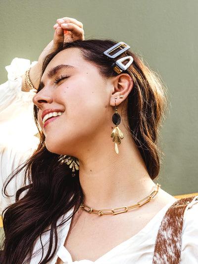 Woman smiling with her eyes closed, wearing two geometric hair clips, gold drop earrings, and a gold chain necklace, styled with a white top.