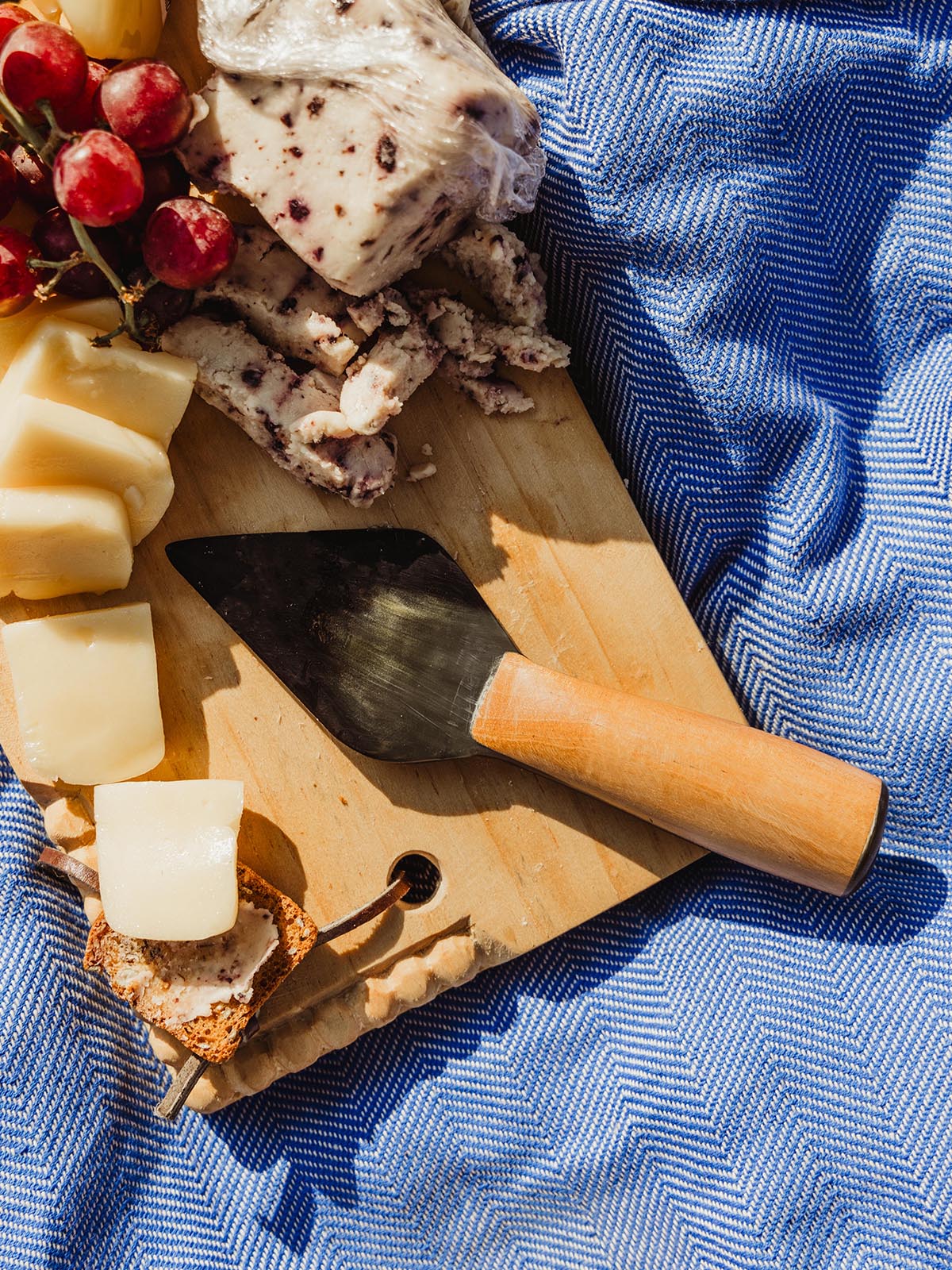 Rustic wooden cheese board with gourmet cheese, crackers, and a handcrafted knife from Joffa, displayed on a blue patterned cloth.