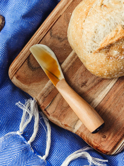 Artisanal butter spreader with a wooden handle on a wooden cutting board next to fresh bread, showcasing Joffa's handcrafted kitchen accessories.