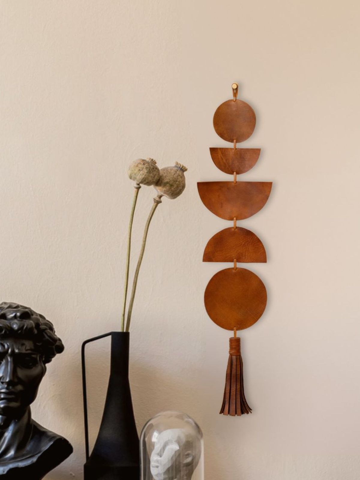 A contemporary interior with a leather geometric wall hanging in earthy brown tones, adorned with a tassel at the bottom. The piece is mounted on a warm-toned beige wall. Below, a black sculptural bust, a modern black vase with dried poppy stems, and a glass cloche covering a minimalist sculpture create a striking contrast of textures and tones.