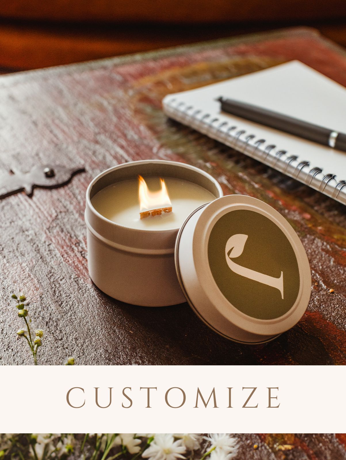 Small candle with company logo. Candle is on a wood surface with a notebook and flowers. Digital sticker to indicate the product is customizable.