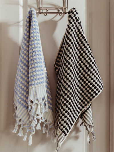 Two woven towels with tassel edges hanging on a metal hook; one features a black and white checkered pattern, and the other has soft blue and white tones.