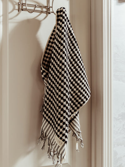 A black and white woven towel with a checkered pattern and striped tassels hanging neatly on a door hook in a well-lit bathroom.