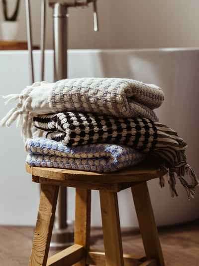 A stack of three textured woven towels in black and white, blue and white, and off-white, folded neatly on a rustic wooden stool beside a modern bathtub.