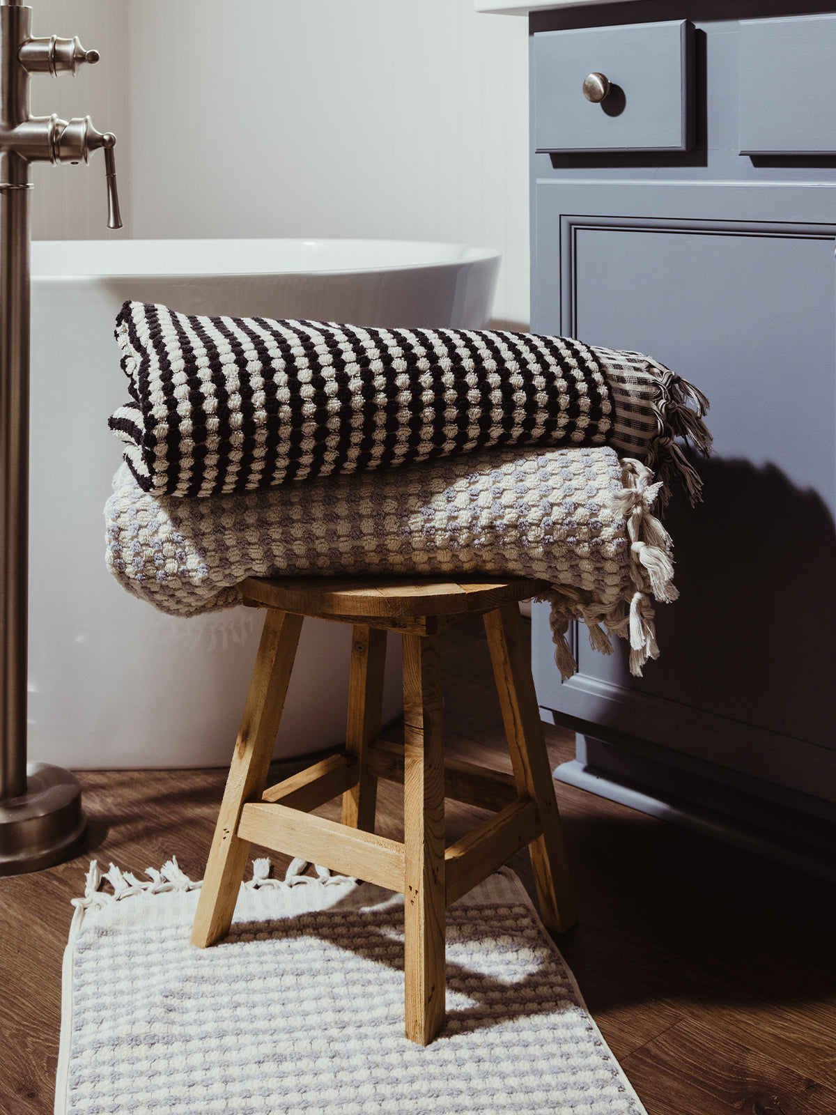 A stack of black and white and grey woven towels with textured patterns and fringe detailing, styled on a wooden stool beside a bathtub for an elegant bathroom aesthetic.