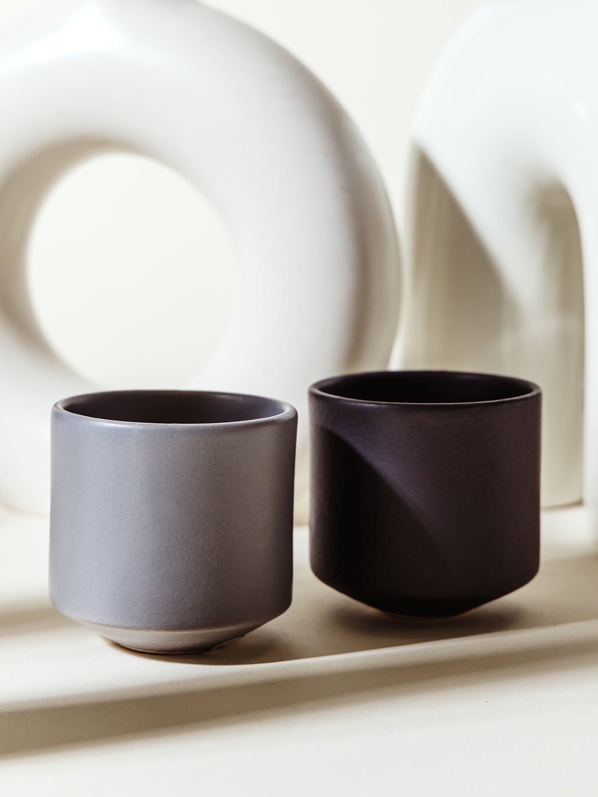 Two modern ceramic cups in muted gray and black tones, set against abstract white ceramic decor. Minimalist and contemporary drinkware perfect for stylish home settings.