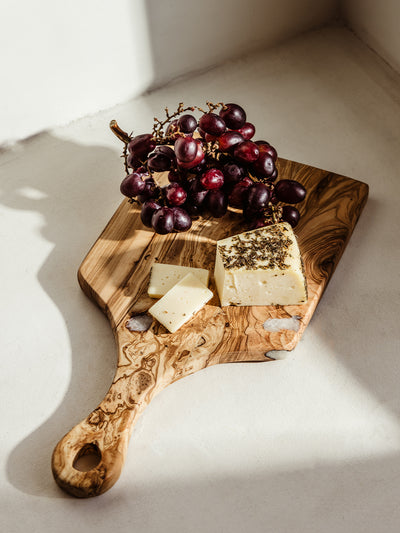 Handcrafted olive wood serving board with gourmet cheese and fresh red grapes, bathed in warm natural light. Perfect for elegant charcuterie presentations and home entertaining.