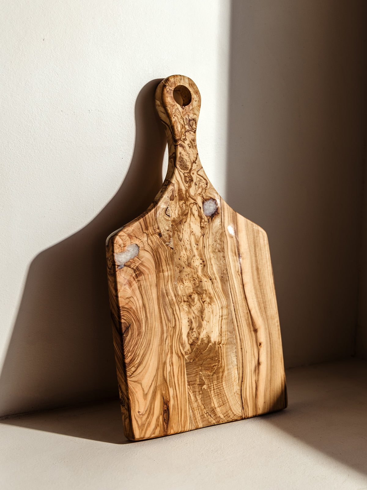 Rustic olive wood cutting board featuring unique grain patterns and natural resin details, leaning against a sunlit wall. A perfect blend of artisan craftsmanship and functionality.