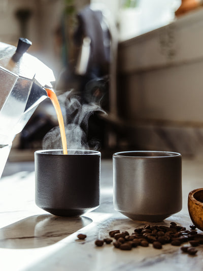 Steaming hot coffee being poured from a stovetop espresso maker into modern matte black and gray ceramic mugs. Coffee beans scattered on a marble countertop add to the artisanal morning aesthetic.