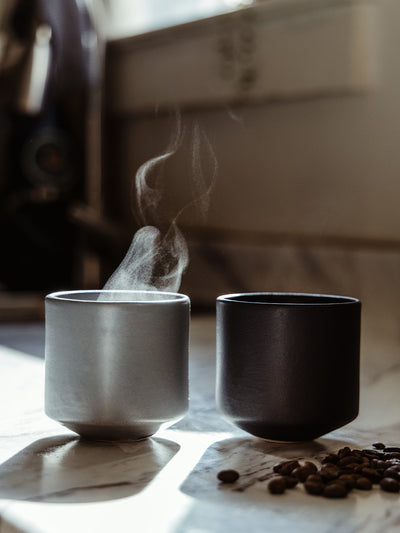 Two minimalist ceramic coffee cups, one black and one gray, with steam rising, set on a marble countertop with scattered coffee beans. A cozy, slow-living moment captured in warm natural light.