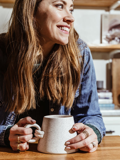 Smiling woman holding a handcrafted speckled ceramic mug with a unique loop handle. A cozy and stylish addition to any morning coffee routine.