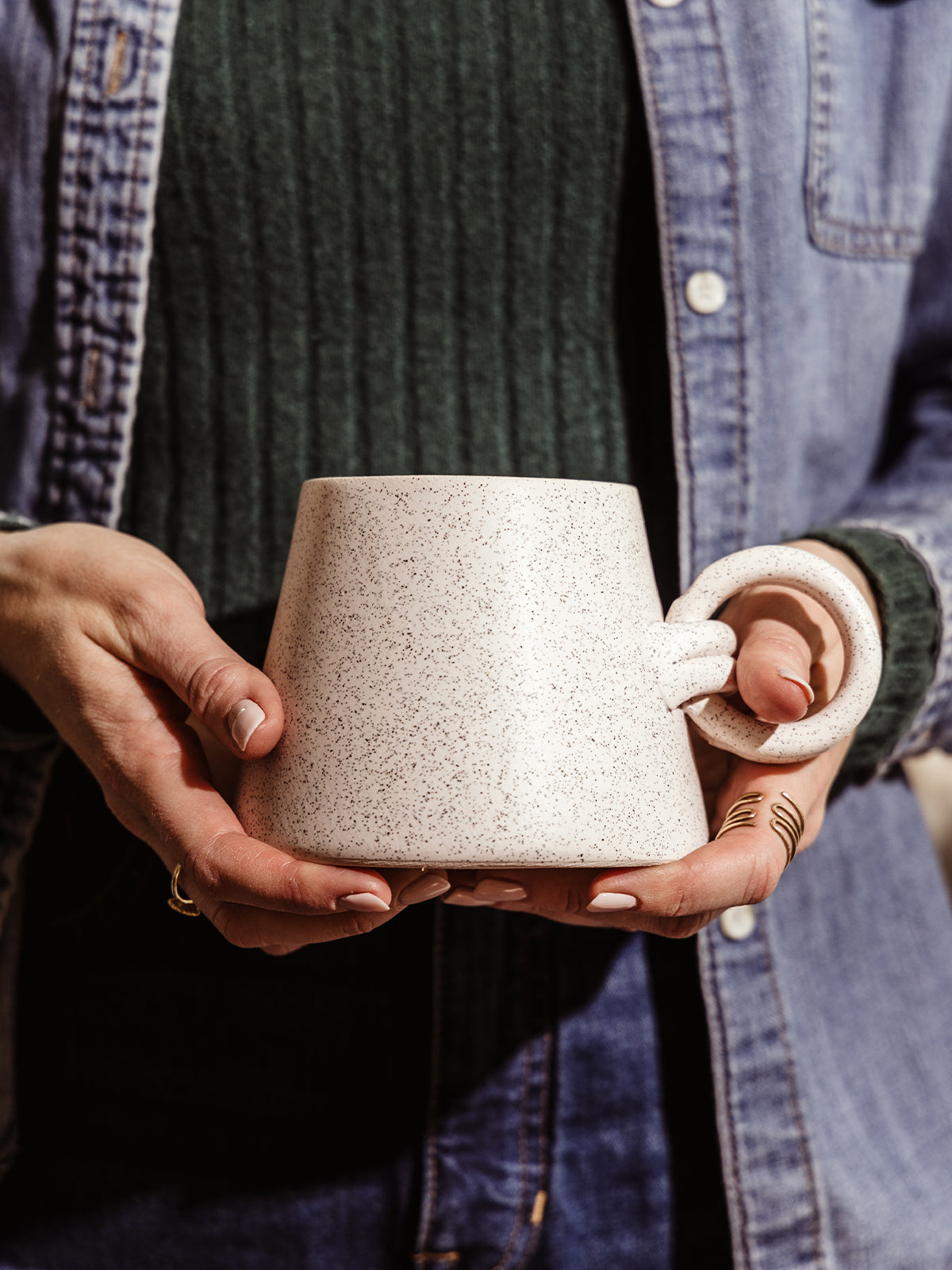 Hands holding a handmade ceramic mug with a speckled glaze and a sculptural loop handle. A minimalist and artistic piece for slow mornings.