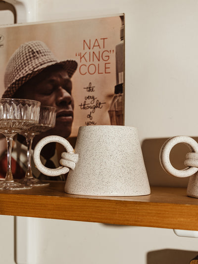 Handmade ceramic mug with a unique knotted handle placed on a wooden shelf, styled alongside vintage decor and glassware.