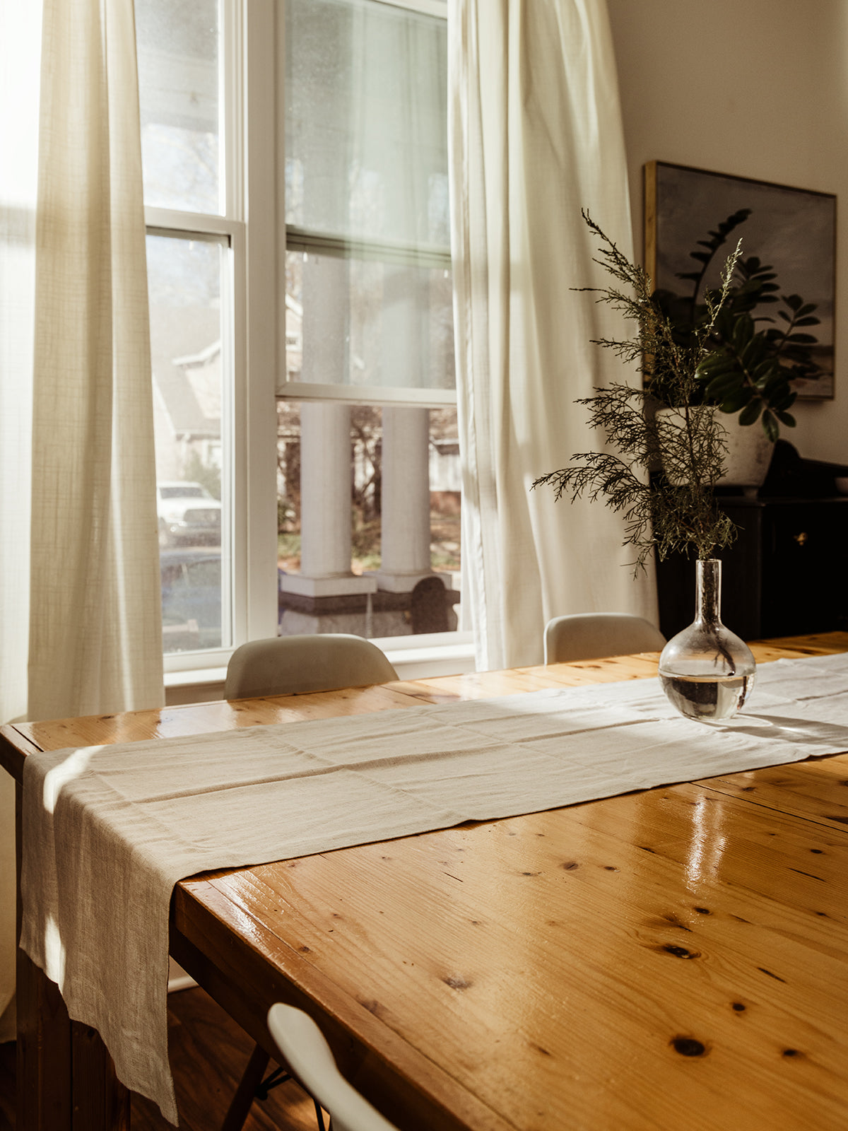 Minimalist dining table styled with a beige linen table runner, a glass vase centerpiece, and natural sunlight streaming through sheer curtains. A simple and elegant home decor touch.