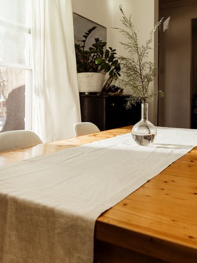 A soft beige linen table runner draped over a rustic wooden dining table, featuring a clear glass vase with delicate greenery for a fresh, minimalist aesthetic.