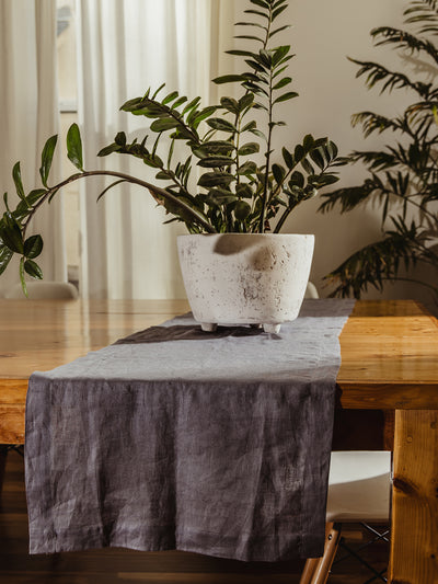 Dining table styled with a gray linen table runner and a rustic white planter with lush green leaves, creating a cozy and modern home aesthetic