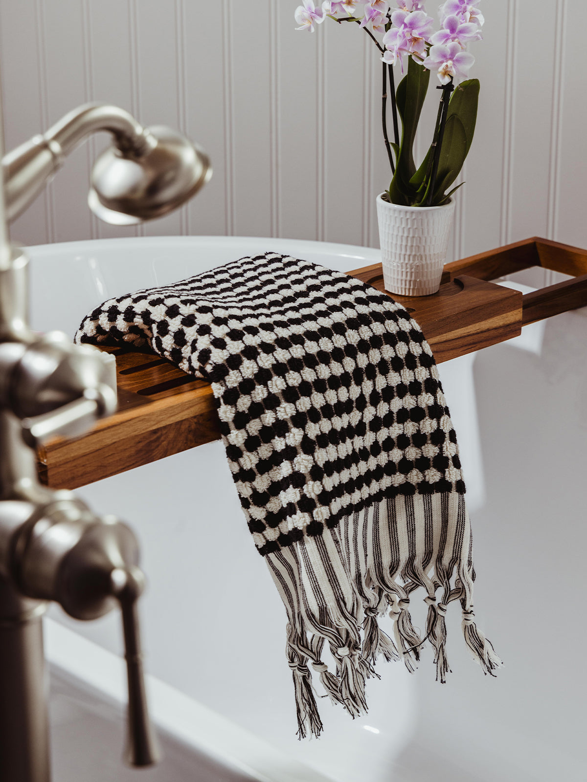 A black and white woven towel with a checkered pattern and fringed edges draped over a wooden bathtub tray, accompanied by a white ceramic pot holding a blooming orchid.