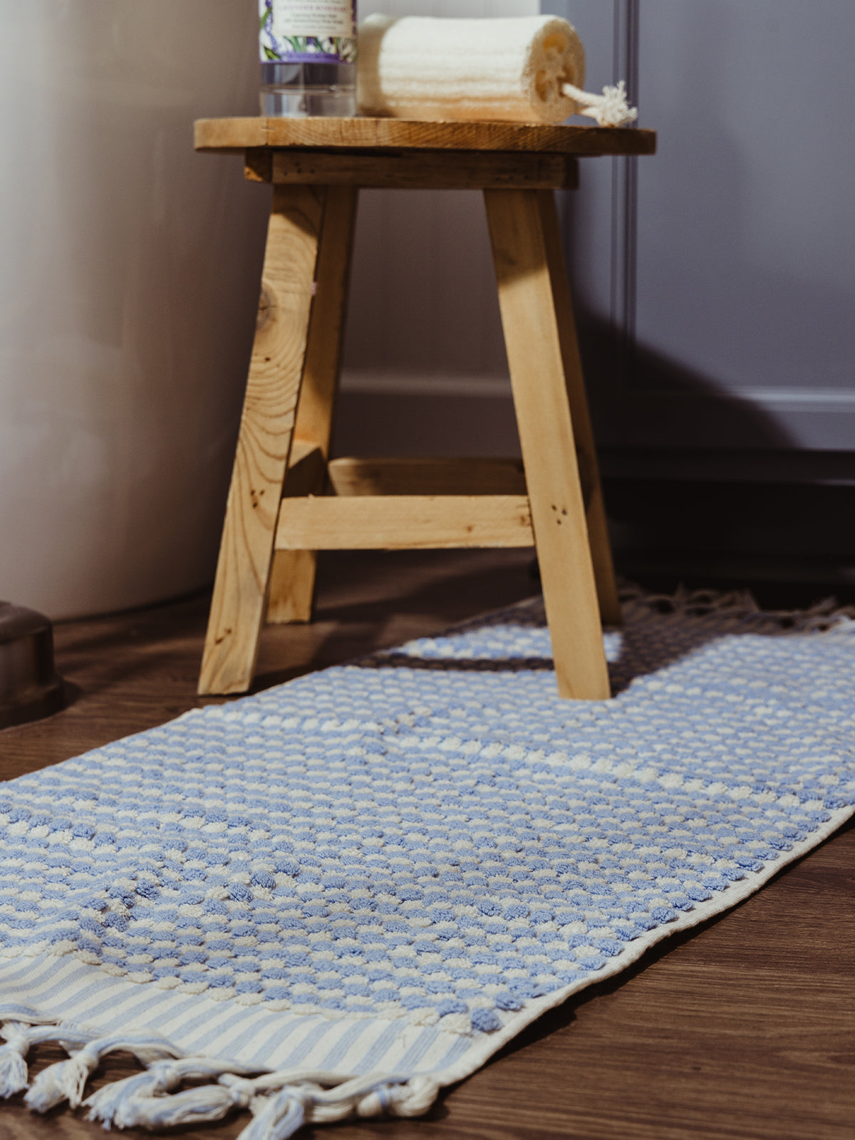 A soft blue and white woven bath mat with tassel edges laid on a wooden floor next to a rustic wooden stool holding a bath sponge and a bottle of bath oil.