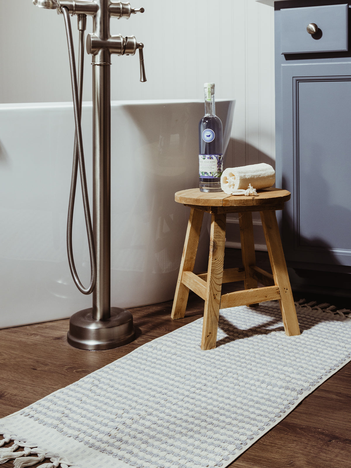 A cozy grey and white bath mat with tassel edges placed in front of a freestanding bathtub, with a wooden stool holding a loofah and a bottle of lavender bath oil.