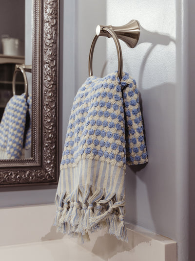 A blue and white striped and textured towel with tassel details hanging on a metal towel ring beside an ornate mirror in a bathroom setting.