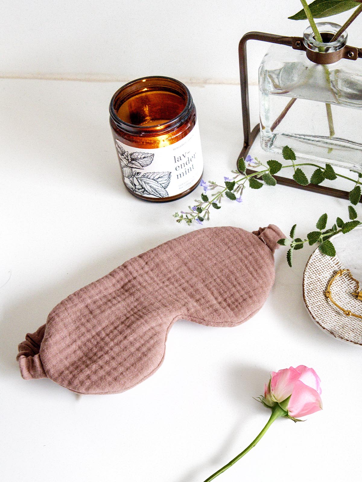 Blush pink fabric sleep mask displayed on a white surface next to a lit candle, a pink rose, and a dish holding gold jewelry, creating a soothing and relaxing setting.