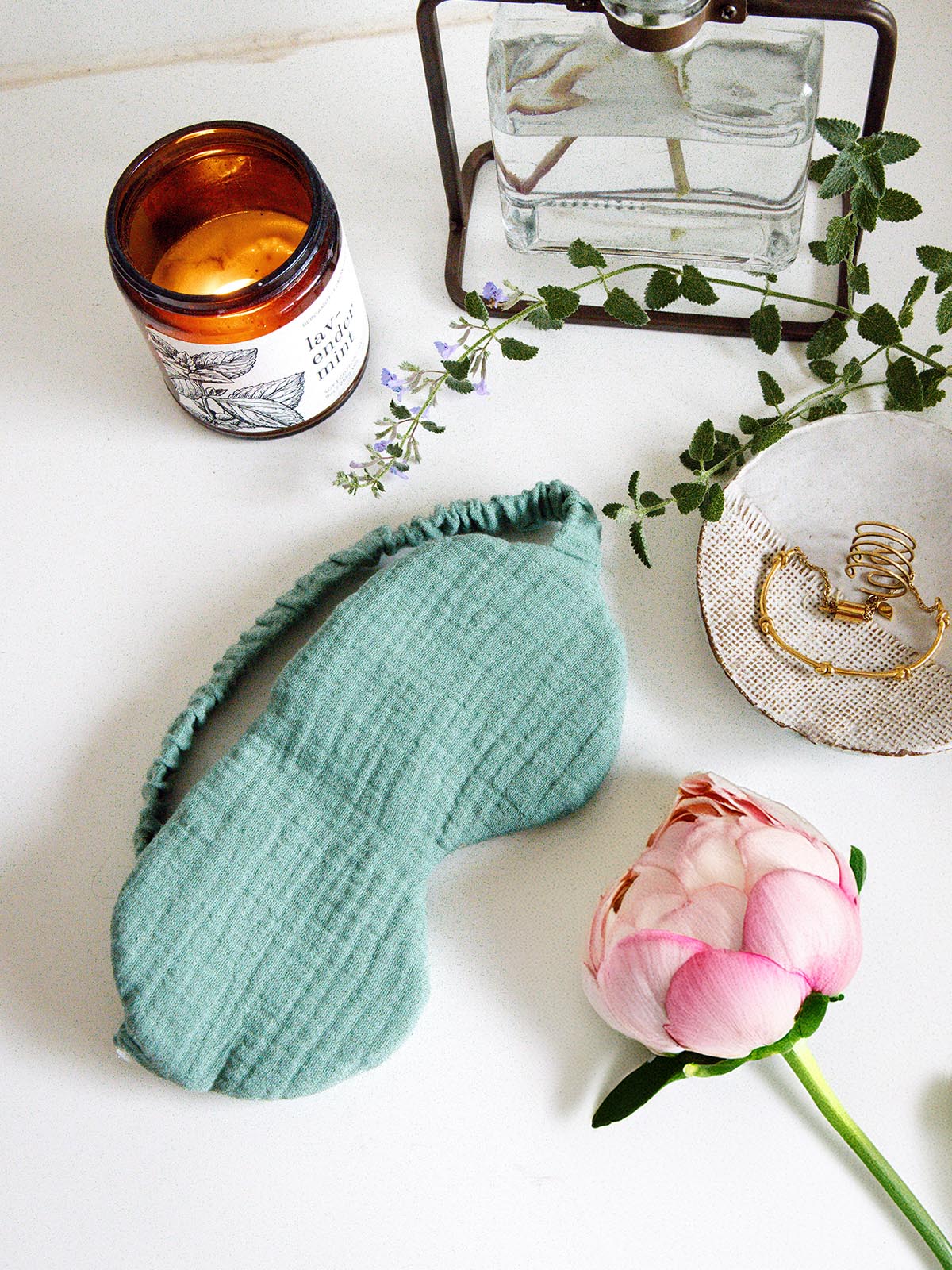 Mint green fabric sleep mask placed on a white surface beside a lit candle, a pink peony, and a dish with gold jewelry, styled to evoke a peaceful and calming atmosphere.