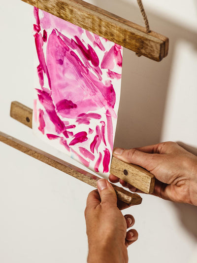 Hands adjusting a pink abstract painting in a wooden art display frame, highlighting the easy-change design.