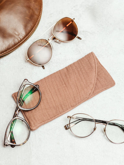 Handmade muslin eyeglass case in soft blush color with eyeglasses and a leather pouch, displayed on a light marble surface.