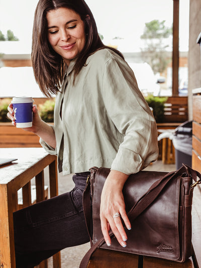 Female model with dark hair sitting at a coffee shop with dark leather satchel. 