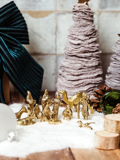 Close-up of a golden miniature nativity scene with handcrafted figurines, set among decorative trees and snow.