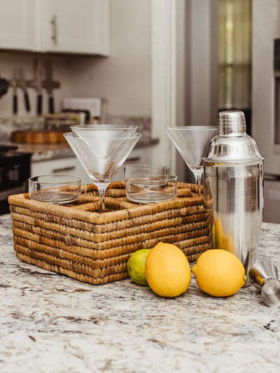 Kitchen countertop with a woven basket holding various cocktail glasses, including martini glasses and coupes. A stainless steel cocktail shaker, lemons, and a lime are placed beside the basket, creating a stylish and ready-to-use drink station.