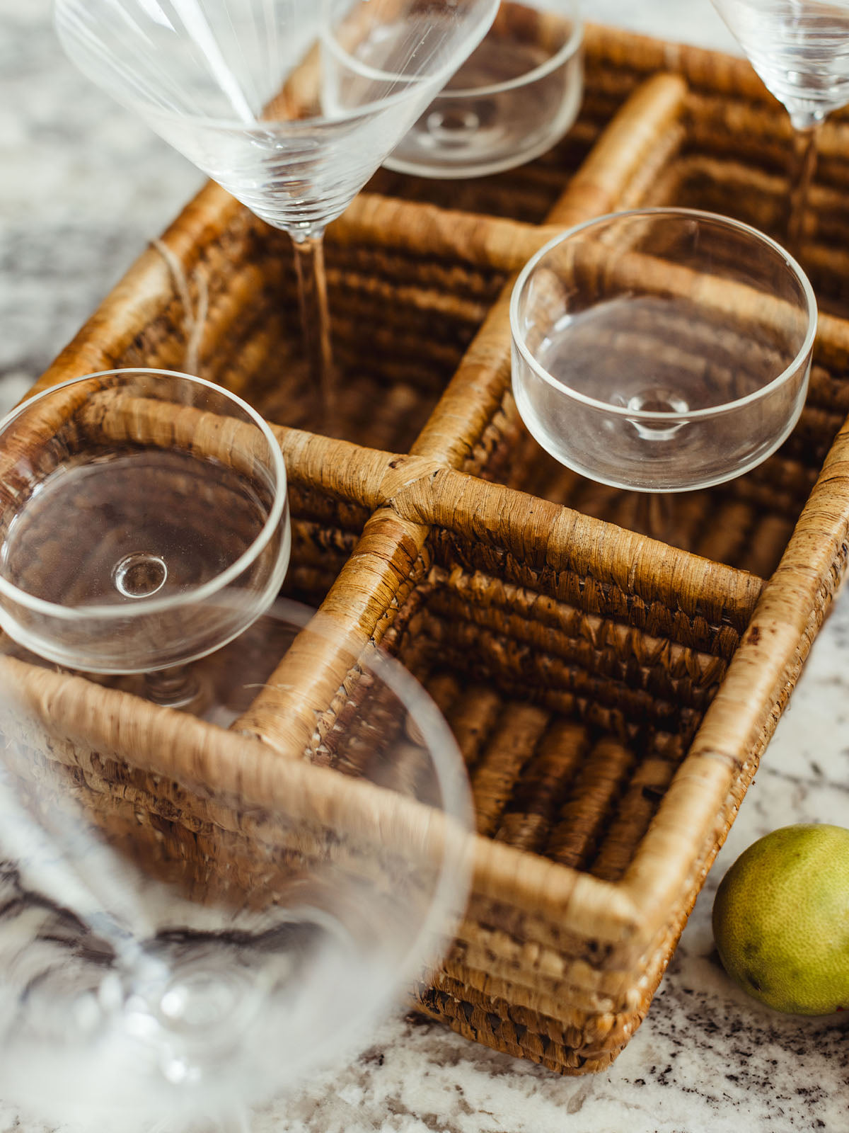 Stylish drink station on a kitchen countertop featuring a woven basket filled with martini glasses and coupes. A stainless steel cocktail shaker, along with fresh lemons and a lime, adds a vibrant touch to the arrangement.