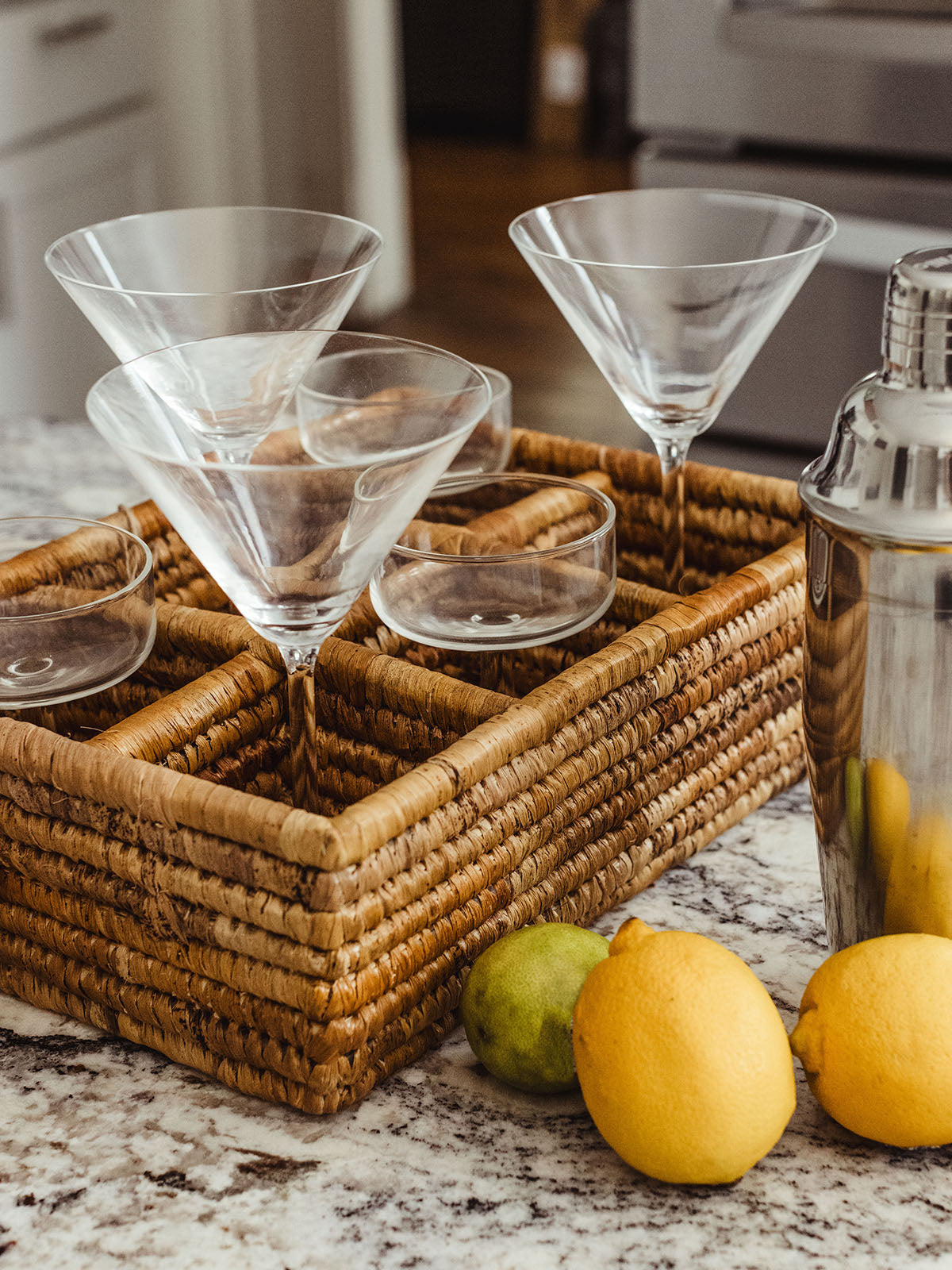 Close-up of a woven basket holding an assortment of cocktail glasses, including martini glasses and coupes. A lime is casually placed next to the basket, emphasizing the rustic yet elegant bar setup.