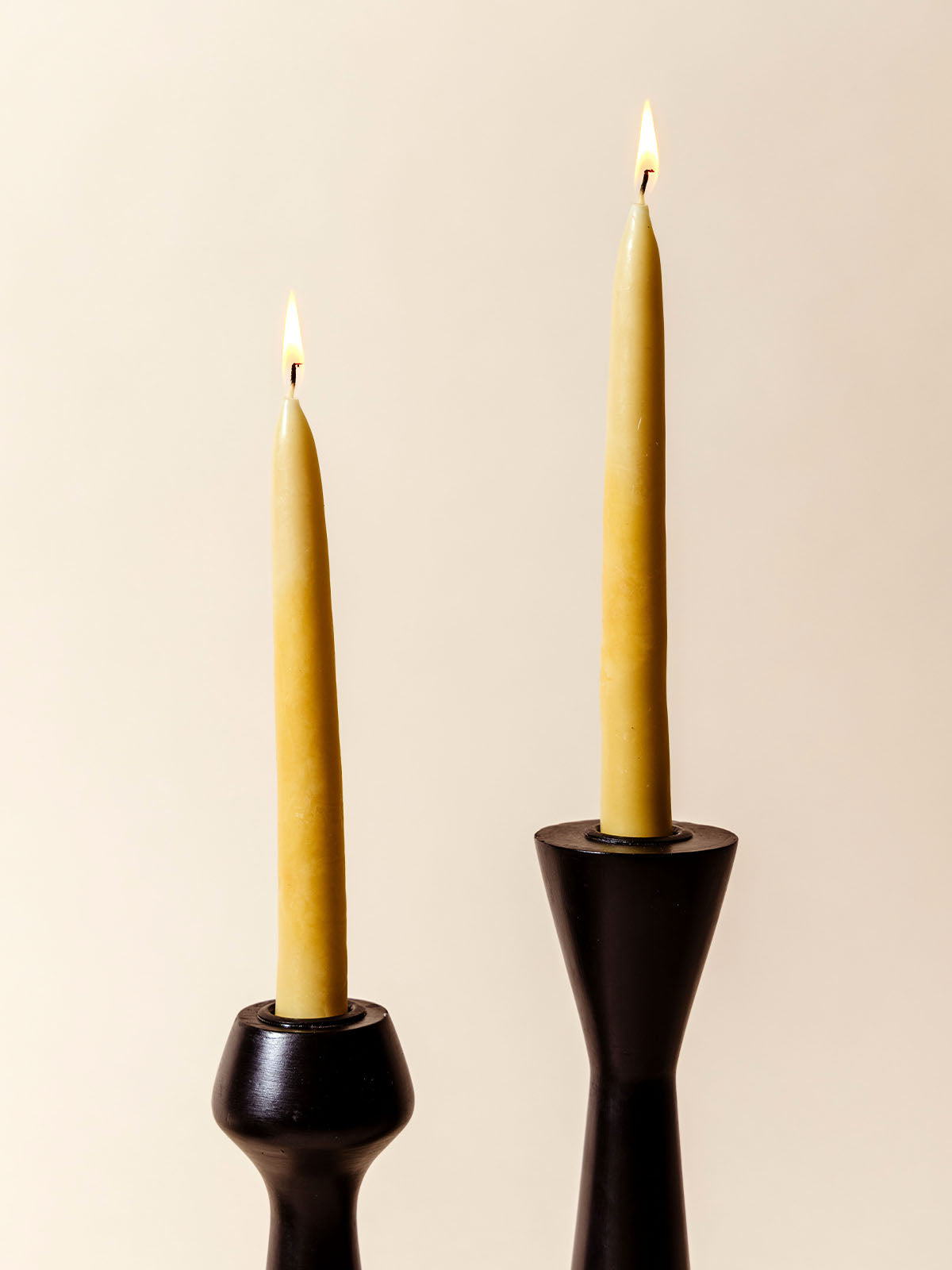 Two lit beeswax taper candles in sleek black candle holders, standing tall against a neutral background. The candles' warm glow contrasts beautifully with the modern, dark holders, creating a minimalist yet elegant ambiance.