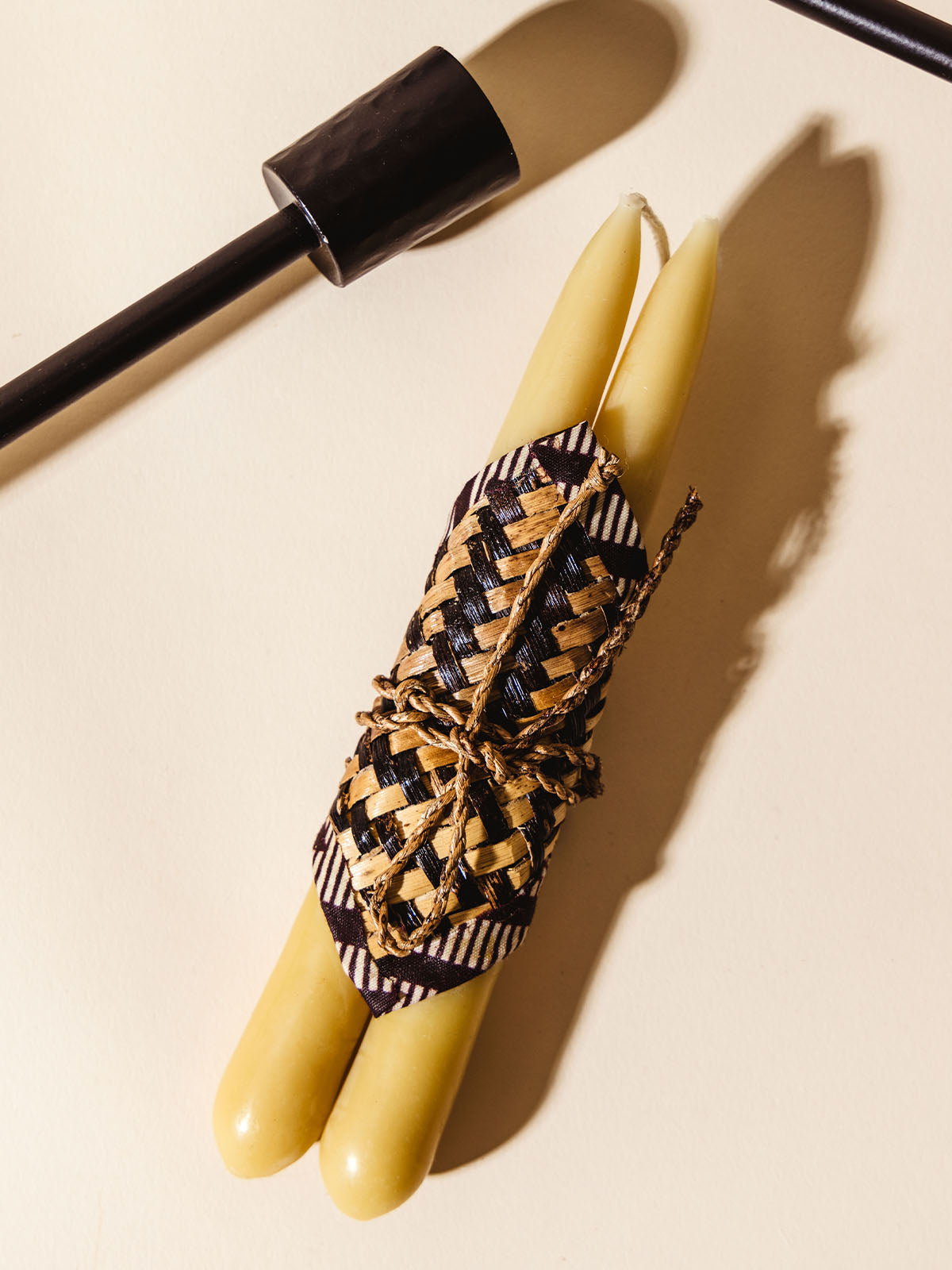 Close-up of two beeswax taper candles wrapped in a decorative woven cover with black and natural-colored stripes. The candles are placed next to a black candle holder, highlighting the artisanal craftsmanship and attention to detail.