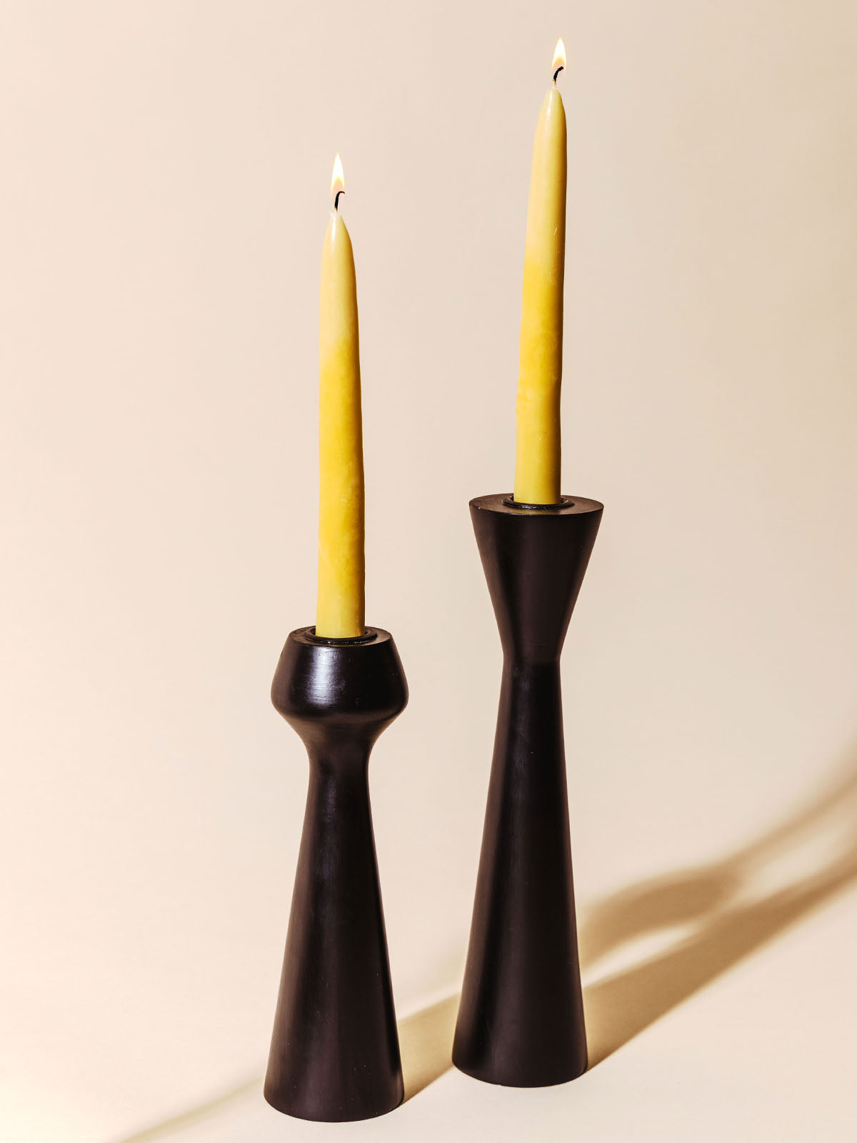 Two lit beeswax taper candles in modern black candle holders, casting soft shadows against a neutral background. The simple, elegant setup showcases the warm light of the candles and the clean lines of the holders.