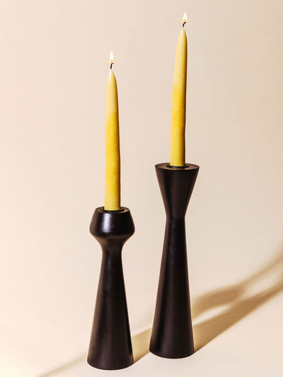 Two lit beeswax taper candles in modern black candle holders, casting soft shadows against a neutral background. The simple, elegant setup showcases the warm light of the candles and the clean lines of the holders.