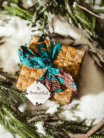 Handwoven wicker gift box with a colorful fabric ribbon, surrounded by frosted evergreen branches on a snow-covered surface, featuring a tag from Beeutiful Creations.