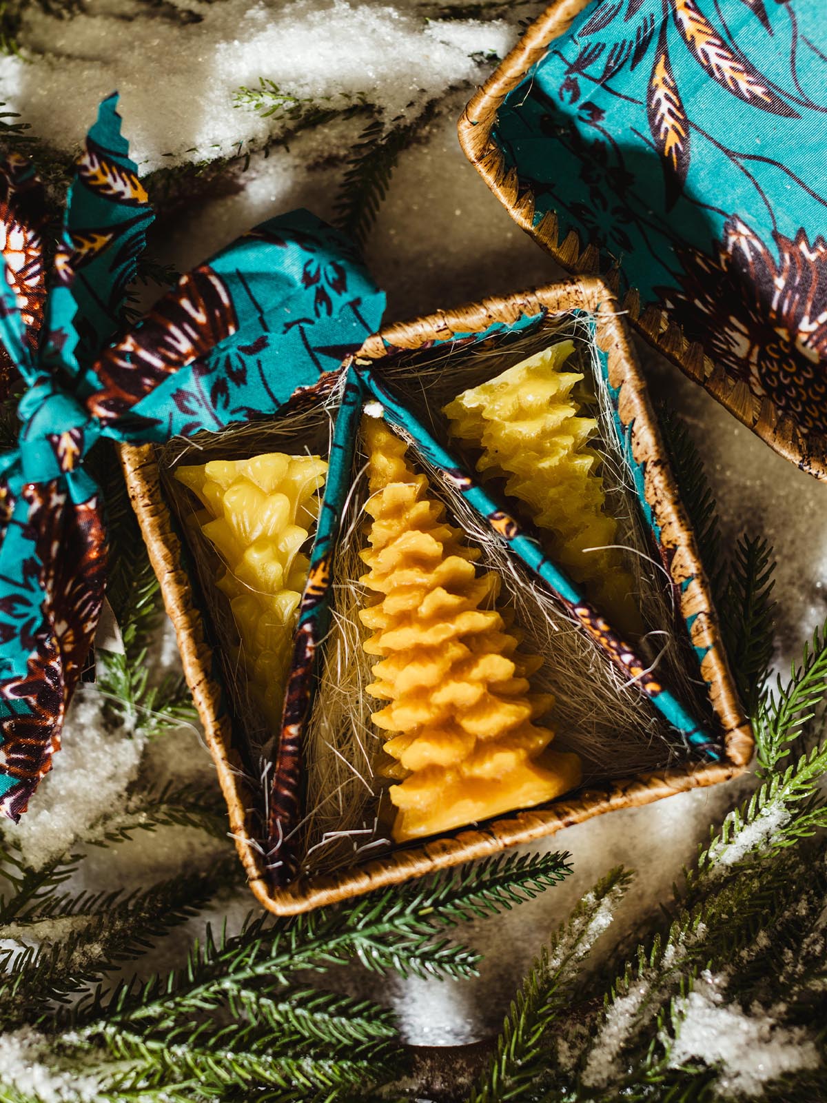 Opened handwoven wicker gift box revealing tree-shaped beeswax candles inside, lined with vibrant fabric and surrounded by frosted evergreen branches on snow.