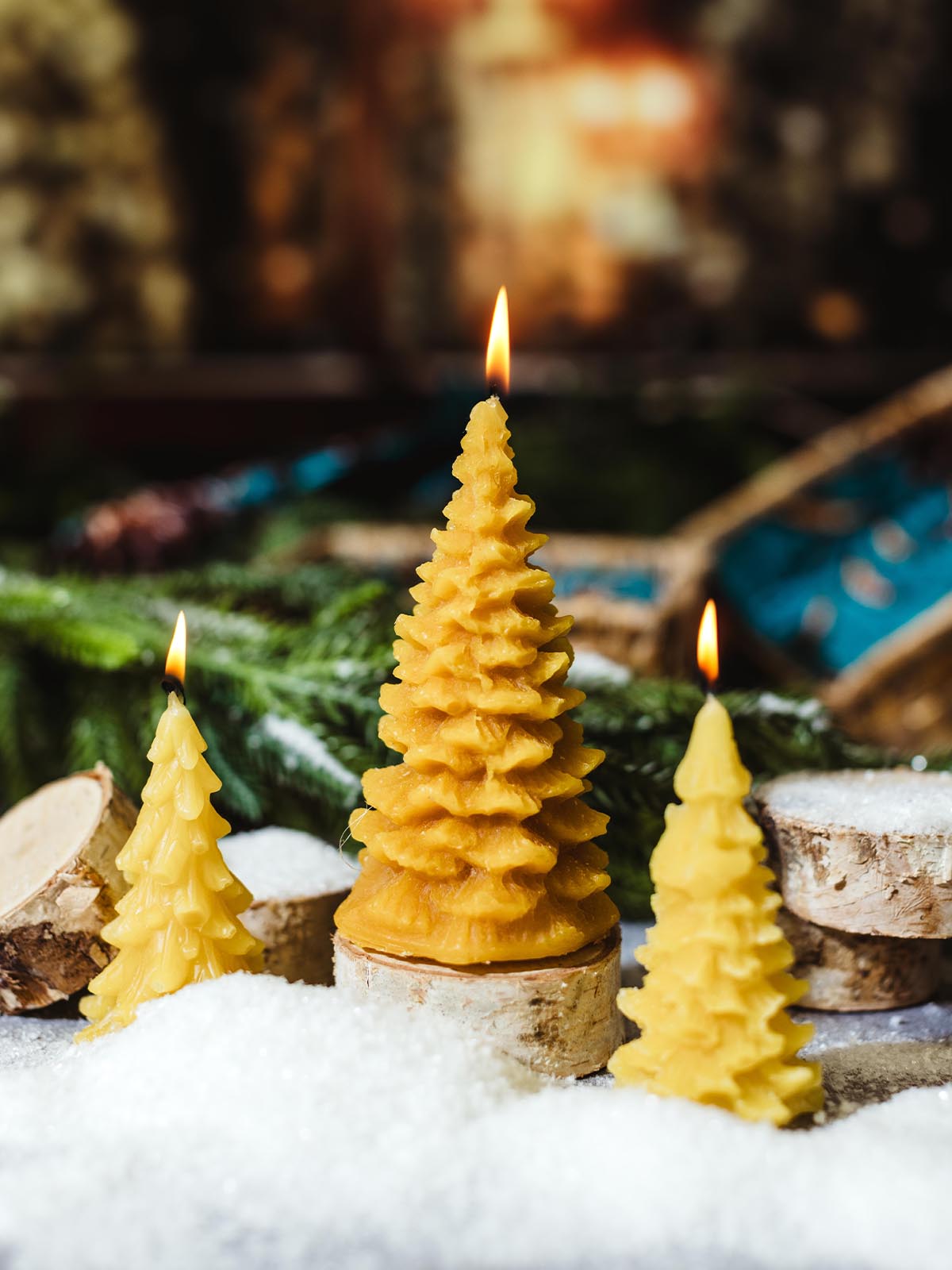 Three tree-shaped beeswax candles in various sizes, burning atop wooden bases in a snowy setting with blurred festive lights in the background.