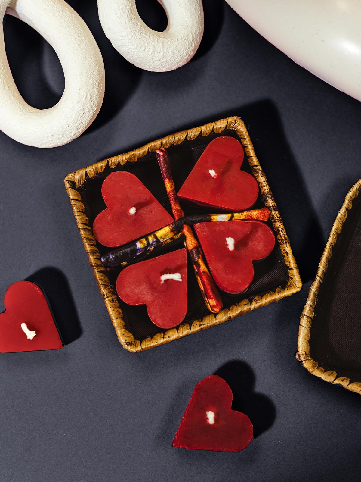 Heart-shaped red beeswax candles arranged in a handwoven wicker box, placed on a black background alongside other decorative elements.