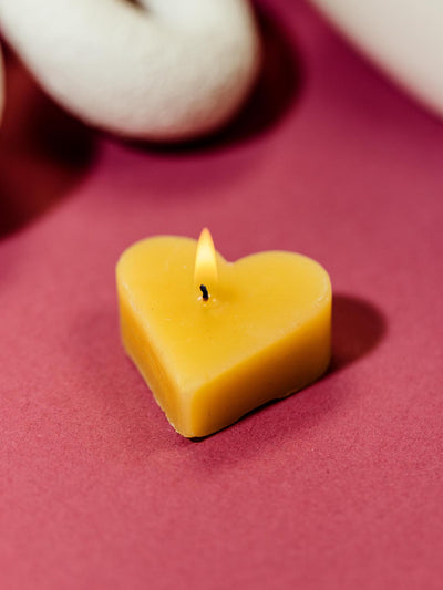 Single yellow heart-shaped beeswax candle burning on a red surface, with white decorative elements in the background.