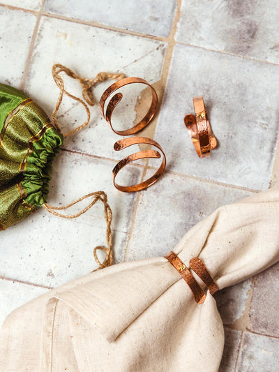 Handcrafted hammered copper napkin rings displayed with a cloth napkin and green drawstring pouch, showcasing the artisanal craftsmanship, available at Joffa.com as part of the sustainable home decor collection.