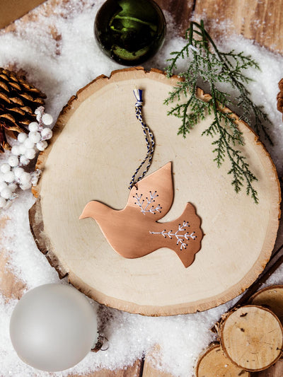 Copper dove ornament with delicate etched details, resting on a wooden slab surrounded by festive greenery and decorations, part of Joffa.com's handcrafted holiday ornament collection.