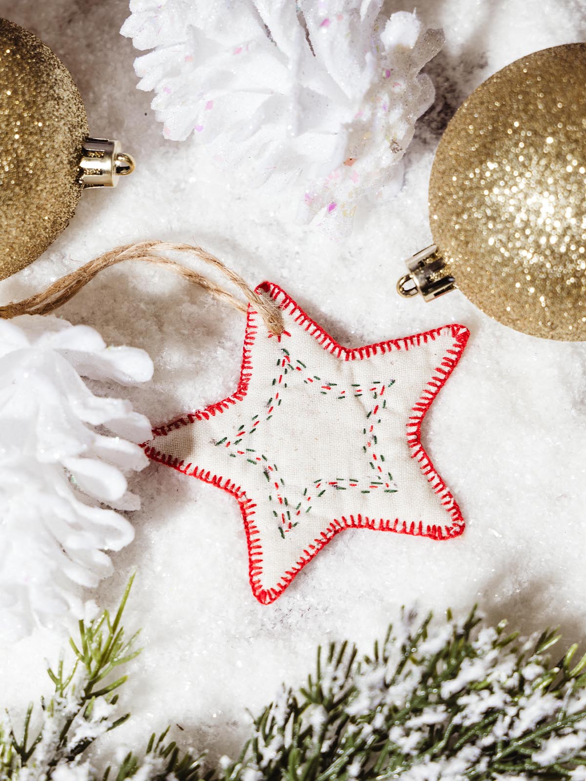 Handcrafted star-shaped ornament with red stitching, resting on snow-like texture surrounded by glittery gold ornaments and white decorative accents. This ethically made holiday ornament adds a festive touch to any decor, perfect for a sustainable Christmas celebration.