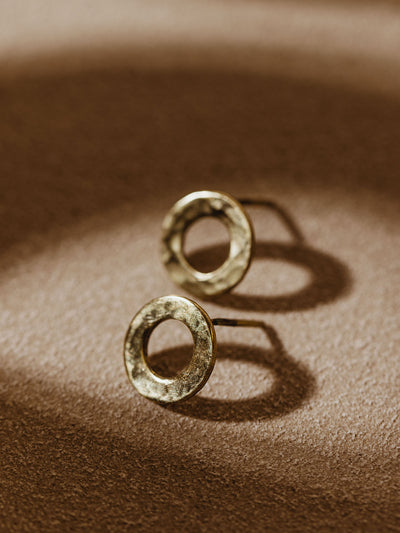 Pair of brass circular stud earrings displayed on a soft, textured surface with warm lighting highlighting their rustic finish.